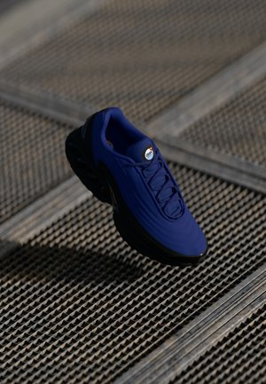 Chaussure de sport bleu foncé avec semelle noire, lacets et logo Air sur la languette, positionnée au-dessus d'une surface métallique texturée avec un motif en grille.