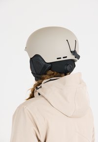 Skihelm in mattem Beige mit schwarzen Akzenten, ausgestattet mit Belüftungsöffnungen und verstellbaren Riemen. Getragen mit einer hellbeigen Jacke.