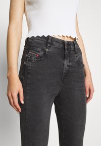 Vaqueros de mezclilla negra de cintura alta con una textura ligeramente desgastada, diseño de cinco bolsillos y herrajes metálicos visibles. Con un detalle de logo rojo.