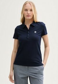 Temno modra polo srajca s kratkimi rokavi, z gumbasto zapiralo in prilegajočim dizajnom. Ima majhen bel logotip na levi prsni strani.