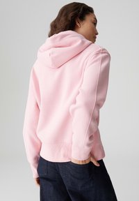 Felpa con cappuccio rosa con finitura testurizzata, cappuccio con coulisse e polsini a costine, abbinata a pantaloni in denim scuro con tasche posteriori.