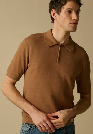 Jeune homme ajustant l'ourlet d'un polo en maille marron à manches courtes, portant un jean bleu, sur un fond beige uni.