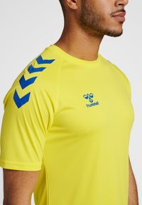 Camiseta atlética amarilla brillante con patrones de chevron azules en los hombros; hecha de un tejido liviano y transpirable con un diseño ajustado.
