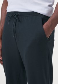Gros plan d'une personne portant un pantalon noir à cordon avec une main dans la poche et une chemise blanche rentrée.