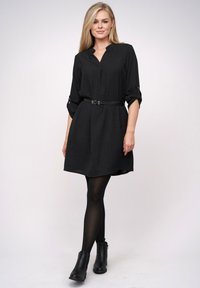 Blonde vrouw in een zwart jurk met riem en opgerolde mouwen, zwarte tights en zwarte enkelboots, staand tegen een effen achtergrond.