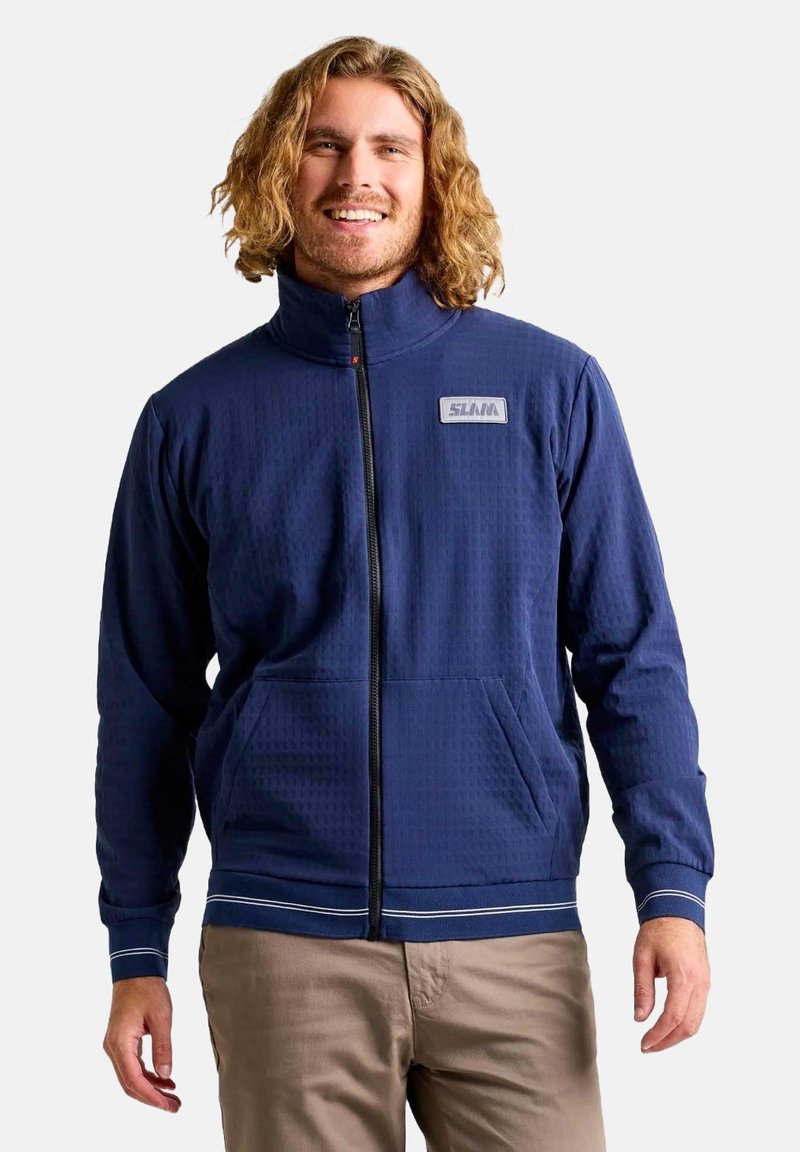 Veste zippée bleu marine en tissu texturé, avec un col montant, deux poches avant et un patch logo sur la poitrine. Rayures blanches en accent au bas.