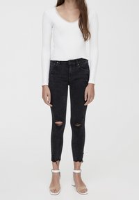 Top blanco de manga larga con un escote adornado con encaje, combinado con unos jeans skinny negros desgastados con dobladillos deshilachados. Sandalias transparentes completan el conjunto.