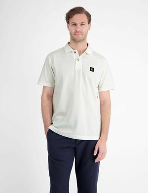 Man staat met handen in de zakken, draagt een wit kortemouw poloshirt met een klein zwart logo en donkerblauwe broek.