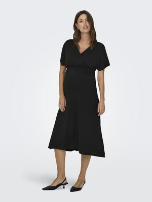 Femme debout portant une robe midi noire à manches courtes et des chaussures slingback noires à petit talon sur un fond clair uni.