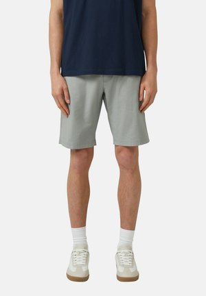 Mann trägt hellgraue knielange Shorts, weiße Socken, beigefarbene Sneakers und ein dunkelblaues Hemd und steht vor einem schlichten Hintergrund.