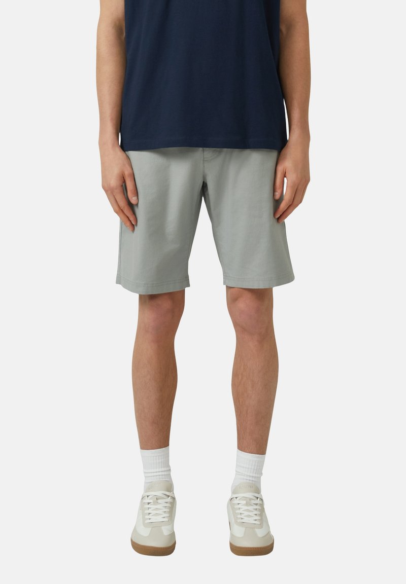 Homme portant un short gris clair jusqu'aux genoux, des chaussettes blanches, des baskets beiges et une chemise bleu marine, debout devant un fond uni.