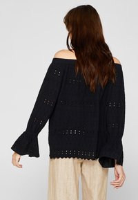 Sort off-the-shoulder bluse med struktureret stof, brede ærmer og udsmykkede mønstre. Har en løs pasform og en diskret flæse ved ærmekanterne.