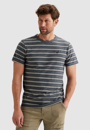 Mann mit kurzen braunen Haaren, der ein dunkelblaues und graues gestreiftes T-Shirt und khakifarbene Hosen trägt, steht mit einer Hand in der Tasche.