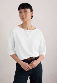 Bílá, volná košile s dlouhými rukávy a širokým výstřihem, v kombinaci s tmavými džíny a pleteným hnědým páskem. Nosené jednoduché zlaté náhrdelníky.