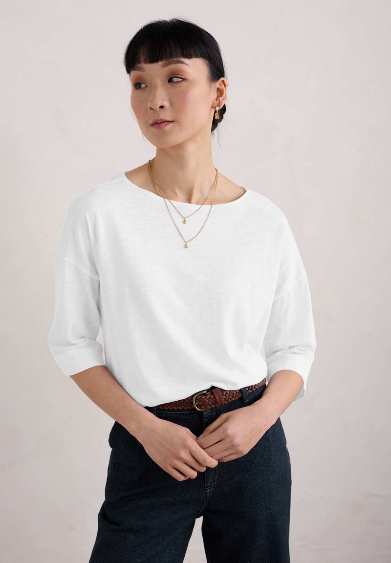 Bílá, volná košile s dlouhými rukávy a širokým výstřihem, v kombinaci s tmavými džíny a pleteným hnědým páskem. Nosené jednoduché zlaté náhrdelníky.