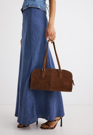 Femme en jupe longue bleue et sandales à talons imprimé léopard tenant un sac à main en daim marron par ses poignées sur fond blanc.