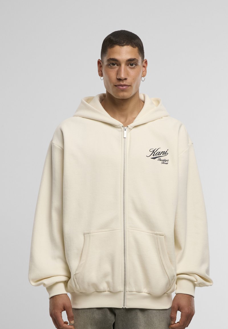 Cream zip-up hoodie with front pockets, a drawstring hood, and embroidered logo on the left side. Soft fabric and relaxed fit.