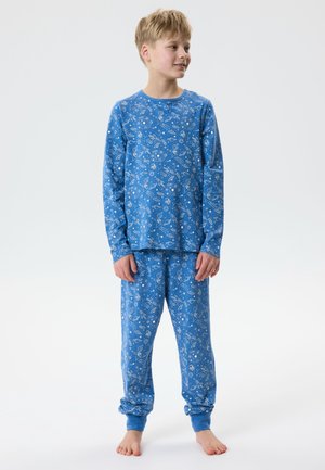 Garçon debout pieds nus portant un haut à manches longues bleu et un pantalon avec des motifs blancs sur le thème de l’espace sur un fond uni.