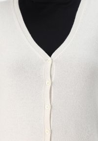 Detailní záběr na krémový svetr s V-výstřihem a čtyřmi knoflíky přes černé tričko.