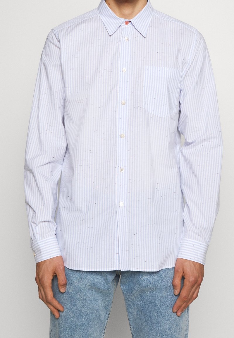 Homme portant une chemise blanche à manches longues avec de fines rayures verticales bleues et un jean bleu clair, debout devant un fond uni.