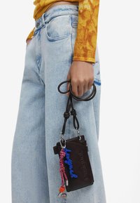 Borsa a tracolla in pelle marrone con dettagli blu e rossi, tracolla appesa e logo "Desigual" in rilievo. Abbinata a jeans a gamba larga blu chiaro.