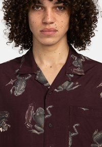Kurzarmshirt in Weinrot aus Stoff mit Froschdruck. Auffällige Details sind eine Tasche und schwarze Knöpfe auf der Vorderseite.
