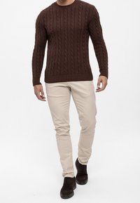 Maglione a trecce marrone, pantaloni slim-fit color crema e stivali scuri. Il maglione presenta un motivo testurizzato e polsini a costine.