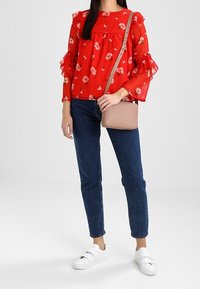 Blusa rossa a fiori con maniche a campana arricciate, abbinata a jeans scuri a vita alta e sneakers bianche senza lacci. Borsa a tracolla beige.