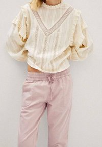 Blusa color crema con mangas rizadas y detalles de encaje; combinada con pantalones de tiro ajustable en un suave tono rosa. Ambas prendas tienen un corte holgado.