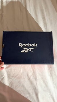 Κουτί παπουτσιών Reebok σε σκούρο μπλε χρώμα με λεία επιφάνεια και λευκό λογότυπο. Διαθέτει καπάκι στην κορυφή και λαβή κομμένη για εύκολη μεταφορά.