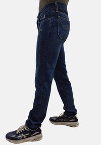 Jeans in denim di colore blu scuro con una vestibilità classica, caratterizzati da cuciture arancioni e cinque tasche, abbinati a sneakers nere.