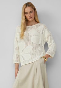 Strickpullover in Creme mit floralem Lochmuster, weitem Ausschnitt und dreiviertelärmeln, kombiniert mit einem beigen Rock.
