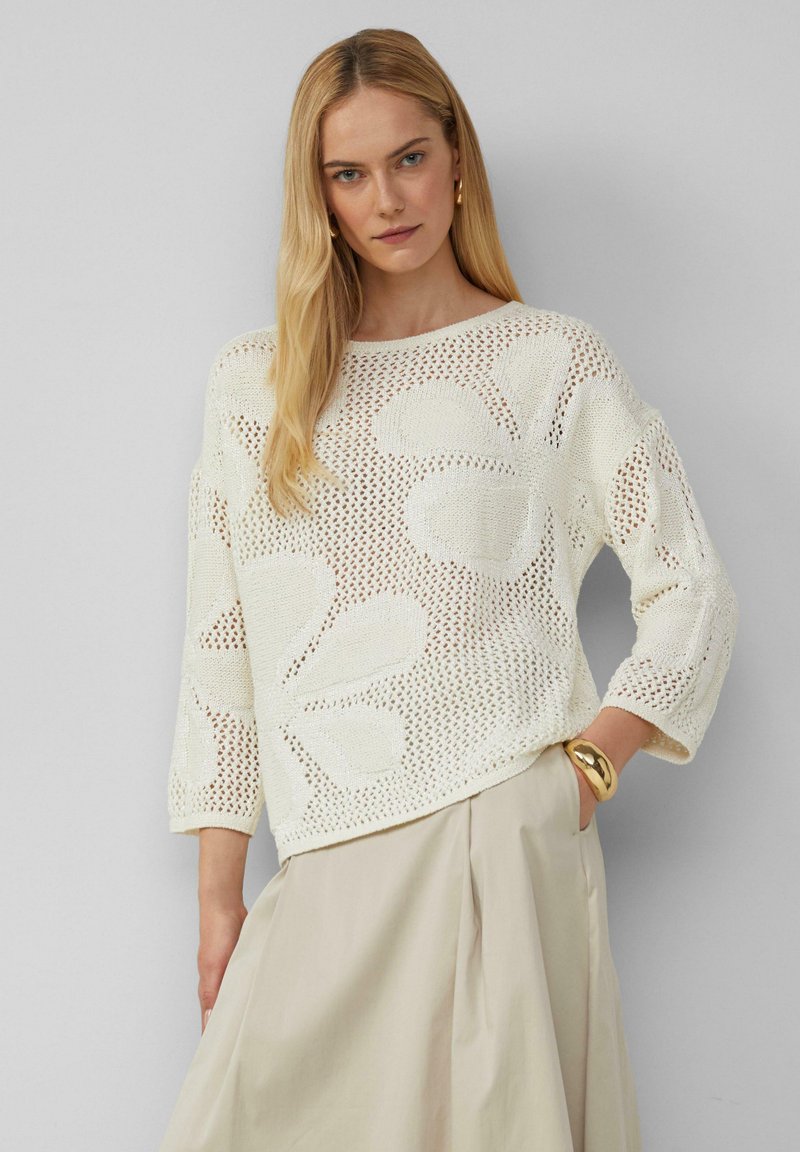 Strickpullover in Creme mit floralem Lochmuster, weitem Ausschnitt und dreiviertelärmeln, kombiniert mit einem beigen Rock.