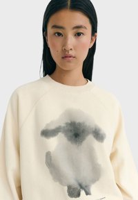 Felpa color crema chiaro realizzata in morbido cotone, con un design di coniglio ad acquerello grigio sul davanti, col collo a girocollo a coste e maniche raglan.