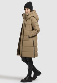 Beige puhvjakot, med hood, med kvilted design, to sidetipper og en sort foring. Kombinert med svarte bukser og ankelstøvler.