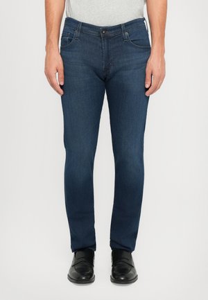 Man wearing slim-fit dark blue jeans and black leather loafers standing against a plain background.