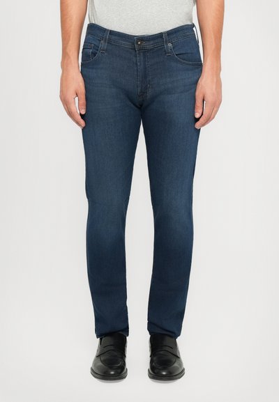 Hombre que lleva jeans ajustados de color azul oscuro y mocasines de cuero negros, de pie frente a un fondo liso.