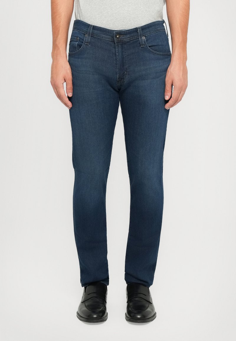 Muškarac u uskim tamnoplave traperice i crnim kožnim polucipelama stoji ispred jednobojne pozadine.