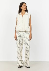 Vesta bej fără mâneci cu fermoar frontal, asociată cu pantaloni ivor cu un model abstract negru, textură fină și o croială lejeră.