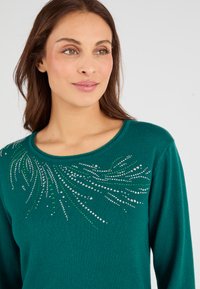 Suéter de color azul verdoso con un escote redondeado, que presenta un diseño decorativo de joyas de plata y verde dispuestas en un patrón fluido.