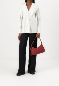 Top bianco a maniche lunghe con un drappo frontale, abbinato a pantaloni neri a gamba larga. Tiene in mano una piccola borsa in pelle color marsala.