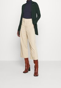 Long green cardigan over a navy turtleneck, paired with beige wide-leg trousers and brown heeled boots. Smooth fabric, tailored fit.