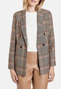 Blazer croisé à carreaux en marron, beige et crème. Caractérisé par une texture lisse, des revers crantés et des poches avant. Associé à un pantalon en similicuir marron clair.
