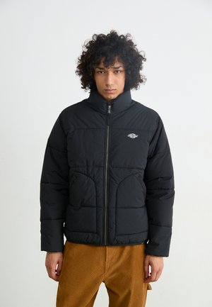 Black puffer jacket with high collar, zip-front closure, and large, quilted panels. Worn over brown corduroy pants.