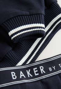 Suéter azul marino con puños acanalados que presentan rayas blancas; cintura elástica etiquetada con "BAKER" en blanco, textura suave, diseño casual.