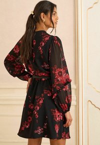 Rochie neagră cu modele florale roșii, mâneci lungi și transparente, manșete volănașe și talie ajustată. Țesătură netedă cu o fustă fluidă.