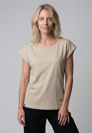Blonde Frau mit schulterlangem Haar, die ein beiges Kurzarmshirt und schwarze Hose trägt, steht vor einem einfarbigen grauen Hintergrund.