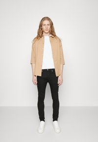 Chemise à manches courtes de couleur bronze sur un t-shirt blanc, associée à un jean noir ajusté et des baskets blanches, se tenant devant un fond uni.