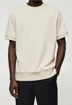 Mørkhudet person iført en ensfarvet, kortærmet beige skjorte og sorte bukser, stående med den ene hånd i lommen mod en neutral baggrund.