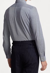 Mężczyzna w jasnoszarej, fakturowanej koszuli z długim rękawem, wpuszczonej w ciemnoniebieskie spodnie w prążki z bocznymi pasami regulacyjnymi, widziany z tyłu.